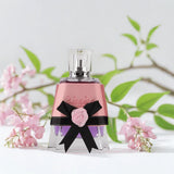 Perfume bottle with pink rose and black bow against a light background with green leaves and pink flowers.